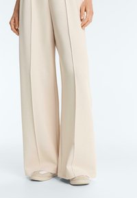 Béžové široké nohavice zakrývajúce béžové a biele slip-on topánky, ruky uvoľnene pri bokoch na jednolutom svetlom pozadí.