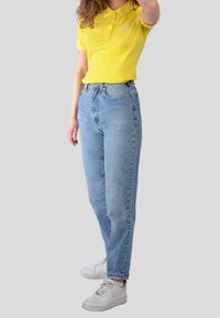 Gul pikétröja med två-knappars placket, tillsammans med ljusblå högmidjade jeans och vita sneakers, som visar en avslappnad outfit.