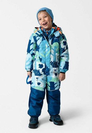 Uśmiechnięte dziecko w niebieskim zimowym kombinezonie w wzory i jasnoniebieskiej czapce, trzymające niebieski hak śnieżny na białym tle.