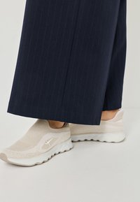 Beige Slip-On Sneaker mit einem strukturierten Strick-Obermaterial und einer klobigen weißen Sohle, kombiniert mit navyblauen, gestreiften weiten Hosen.