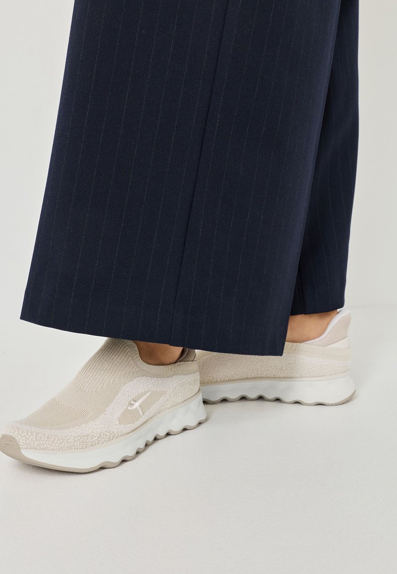 Beige Slip-On Sneaker mit einem strukturierten Strick-Obermaterial und einer klobigen weißen Sohle, kombiniert mit navyblauen, gestreiften weiten Hosen.