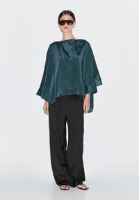 Teal satin ponchotopp med asymmetrisk fåll, kombinerad med löst sittande svarta byxor och platta sandaler. Modellen bär stora mörka solglasögon.