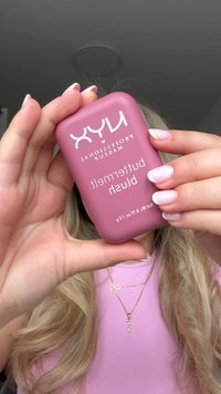 NYX Rouge in einem abgerundeten, sanft rosa kompakten Behälter. Weißer Text zeigt Produktname und Beschreibung. Glatte Textur; Kompakt wird zwischen den Fingern gehalten.