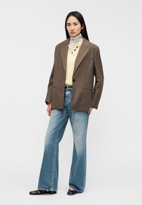 Blazer marrón a medida sobre un suéter crema con cuello a rayas, combinado con jeans acampanados azules descoloridos y mocasines negros.