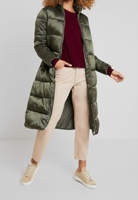 Lungo cappotto imbottito verde oliva, con zip frontale e colletto alto. Indossato sopra un maglione bordeaux, abbinato a pantaloni beige e sneakers chiare.