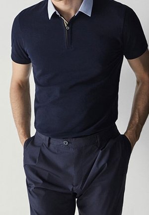 Mann trägt ein marineblaues kurzärmeliges Poloshirt mit hellblauem Kragen und dunkelblauer Hose, Hände in den Taschen.