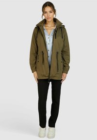 Olivgrüne Zip-Jacke mit Kapuze, verstellbaren Kordeln, Seitentaschen und lockerer Passform, kombiniert mit schwarzen Hosen und weißen Sneakern.