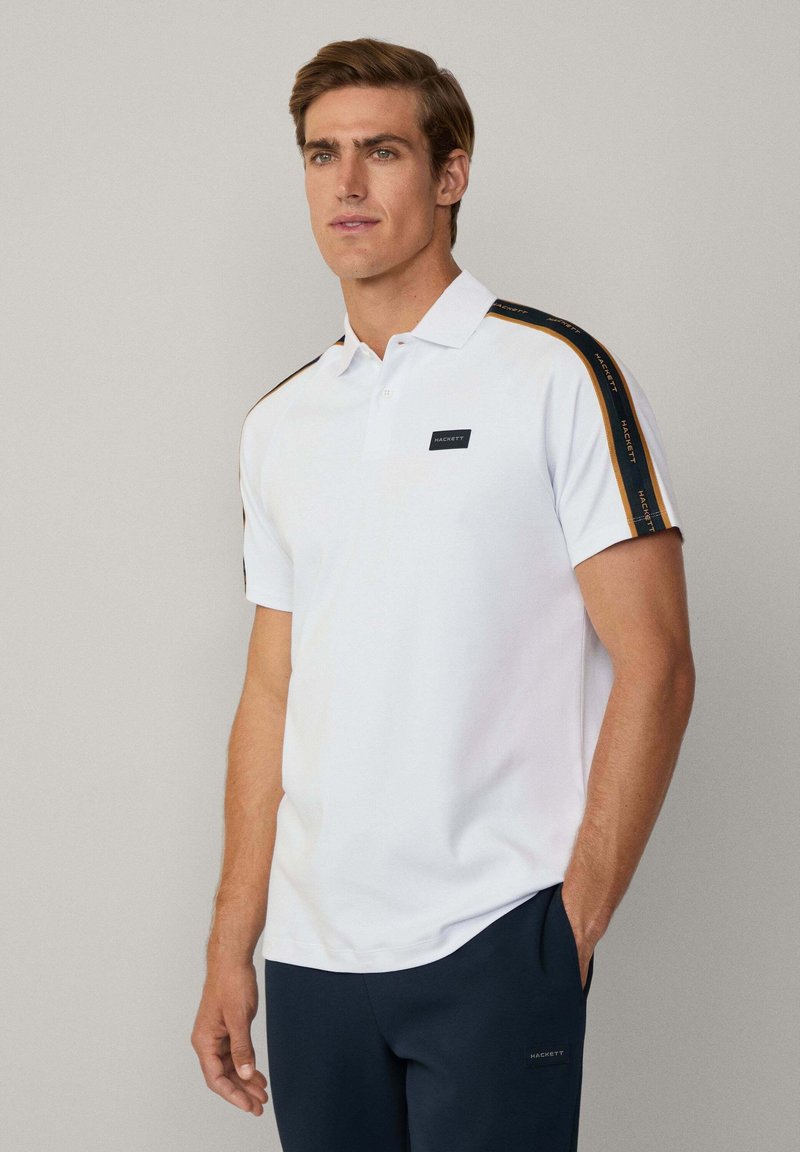 Hvid polo shirt med krave, korte ærmer, sorte og gyldne sideløsninger, og et lille sort logo-patch på brystet. Glat stofstruktur.