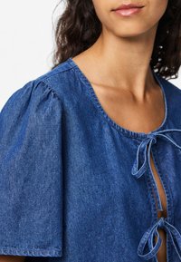 Τζιν τοπ με κοντά φουσκωτά μανίκια, βαθύ λαιμόκομμα και κλείσιμο με κορδόνι μπροστά. Το ύφασμα είναι απαλό, σε μέτριο μπλε χρώμα με μία διακριτική υφή.