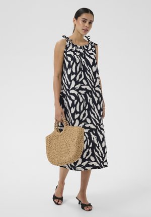 Femme portant une robe midi sans manches à motif noir et blanc avec des bretelles nouées aux épaules, tenant un sac à main tressé beige et portant des talons ouverts noirs.