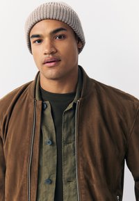Giacca bomber marrone sopra una camicia verde, con bottoni blu; abbinata a un berretto a coste color beige chiaro. Tessuto dalla texture liscia, design casual.