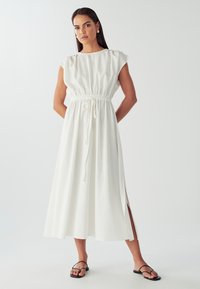 Bílé šaty z lněné směsi s volným střihem, krátkými cap-sleeve rukávy, zavazováním v pase, bočními rozparky a délkou ke kotníkům. Černé sandály.