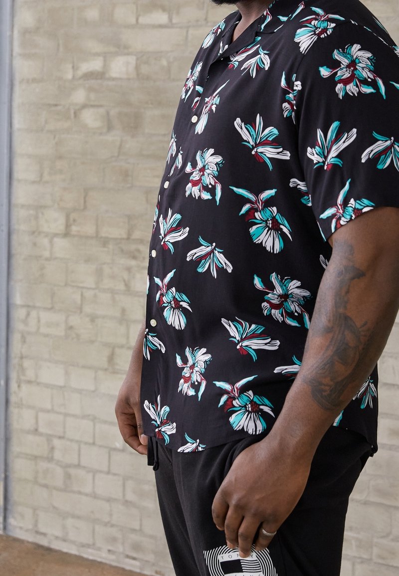 Homme portant une chemise noire à manches courtes boutonnée avec un imprimé floral bleu et blanc, debout devant un mur en briques clair.