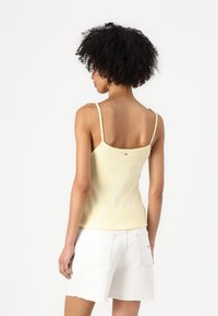 Kollane ribaga tank-top õhukeste rihmadega ja lainelise äärisega, kantud valgete denimpükstega, millel on tagakotid. Tagaküljel on logodetail.