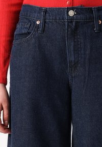 Jean en denim foncé avec une coupe droite, dotés de poches avant, de rivets en cuivre et d'un détail de couture visible sur la jambe. Associé à une chemise rouge.