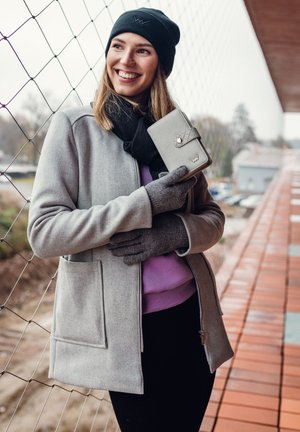 Smiling woman in gray coat, black hat, gloves, and scarf holds gray wallet against a balcony railing with netting outdoors.