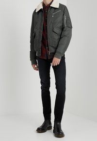 Giubbotto bomber verde oliva con colletto in pelliccia beige, tasche con zip, polsini a costine, indossato sopra una camicia a scacchi rossi e neri, jeans neri, stivali neri.