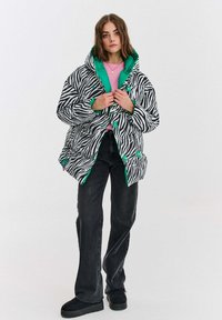 Žena s vlasmi po plecia, so svetlo hnedými vlasmi, oblečená v pufrovej bunde so zebrovým vzorom v čierno-bielej kombinácii s zeleným podšívkami, ružovom topom, čiernych džínsoch a čiernych topánkach.