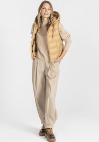 Beżowa kamizelka puchowa z kapturem, założona na jasny sweter, w połączeniu z szerokimi spodniami w kolorze kremowym i brązowymi sznurowanymi butami na grubej podeszwie.