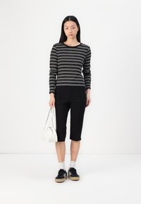 Randig långärmad topp i svart och vitt, kombinerad med svarta capribyxor. Modellen håller en liten vit väska, bär svarta sneakers och vita strumpor.