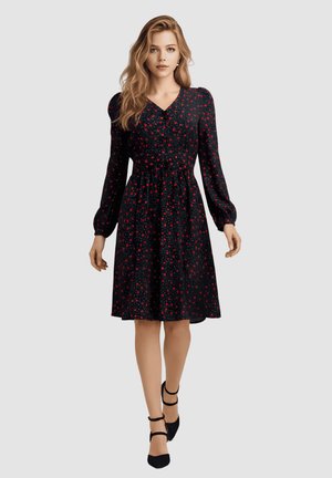 Mujer con vestido negro hasta la rodilla con estampados florales rojos y mangas largas, caminando hacia adelante con zapatos de tacón de correa en el tobillo negros sobre un fondo gris.