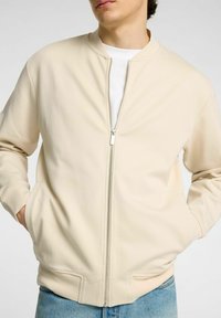 Hombre vestido con una chaqueta cremosa con cremallera sobre una camisa blanca, con las manos en los bolsillos de la chaqueta, combinado con jeans azules, contra un fondo sencillo.