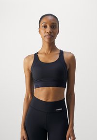Black sports bra with a round neckline and wide straps, made of stretchy fabric. Features a smooth texture and a logo on the waistband.