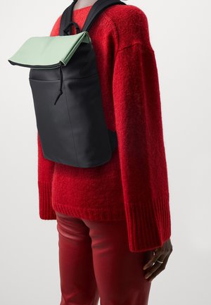 Musta reppu, jossa on taitettu mintunvihreä läppä, valmistettu sileästä materiaalista ja varustettu säädettävillä mustilla hihnoilla. Puettuna punaisen kuvioidun neuleen päälle.