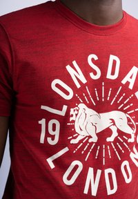 Rød mønstret t-shirt med rund hals, der har et hvidt løvegrafik og teksten "Lonsdale London 19" med fede bogstaver.