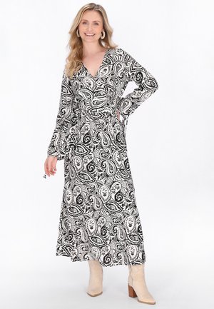 Smilende kvinde iført lang sort og hvid kjole med paisleymønster og lange ærmer samt beige støvler med hæl, stående med den ene hånd på hoften.