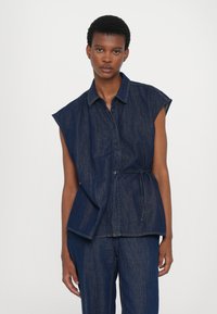 Camisa sem mangas de denim escuro, com frente abotoada, colarinho, cintura ajustada e fendas laterais. Tecido texturizado com variações subtis na tonalidade.