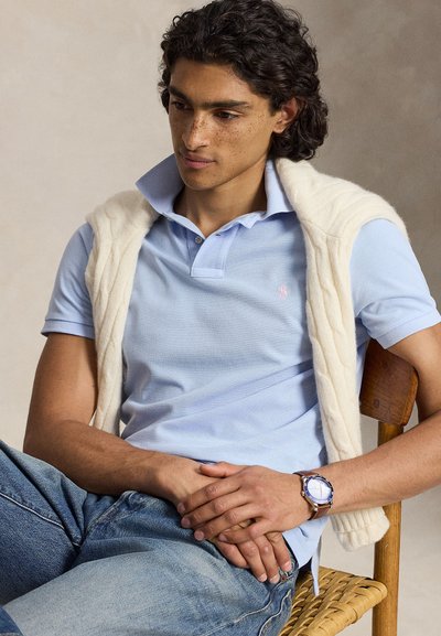Jeune homme aux cheveux bouclés portant un polo bleu clair, un pull crème drapé sur les épaules, un jean bleu, une montre en cuir marron, assis sur une chaise en bois.