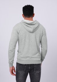 Man with short curly hair wearing a light gray hoodie and dark washed jeans, standing facing away against a plain light background.