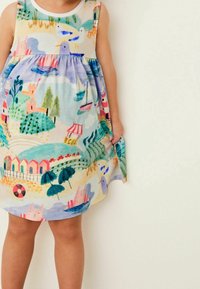 Dítě v barevných šatech s ilustracemi lodí, ptáků, stromů, stánku s zmrzlinou a plážovými slunečníky, stojí před jednobarevným pozadím.