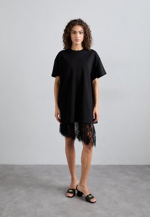 Fekete, oversized póló, puha pamutból, rövid ujjakkal és csipkés szegéllyel. Fekete csúszó szandálokkal viselve.