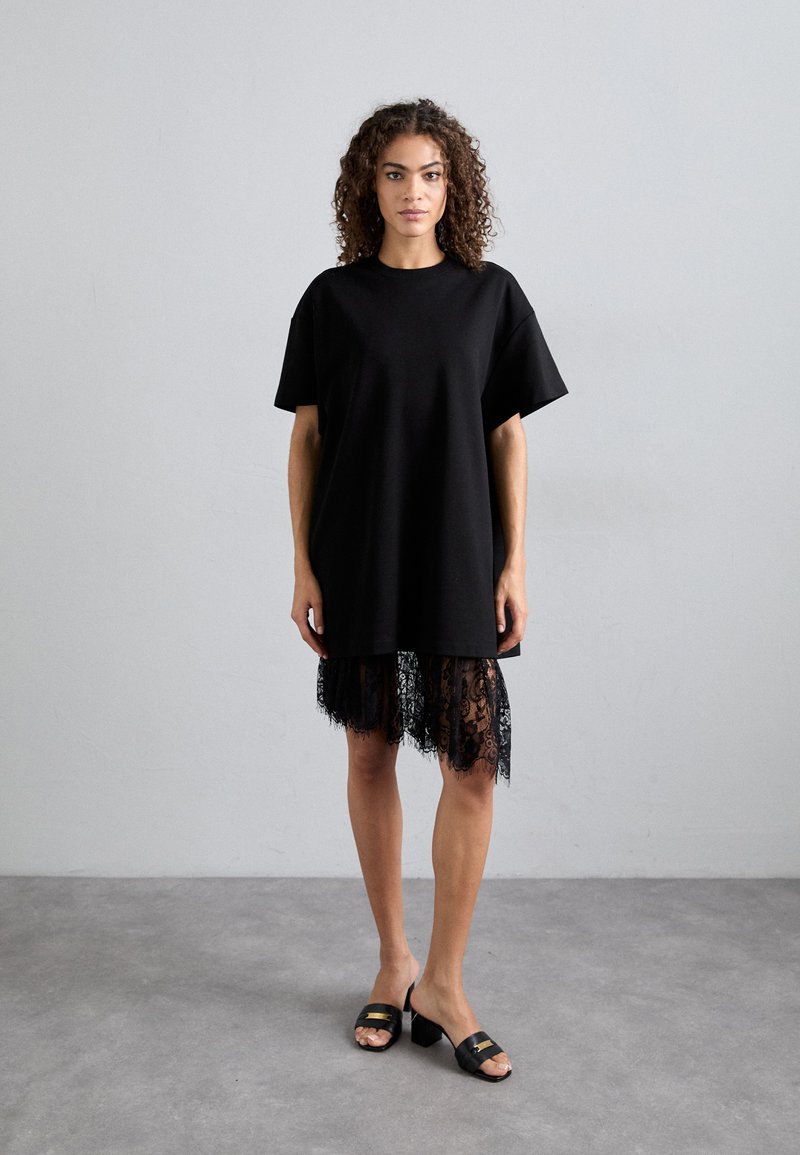 Fekete, oversized póló, puha pamutból, rövid ujjakkal és csipkés szegéllyel. Fekete csúszó szandálokkal viselve.