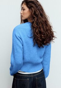 Hellblauer Strickpullover mit rundem Ausschnitt und geripptem Saum. Das Modell trägt ihn über einem weißen Top, kombiniert mit dunklen Denim-Jeans.