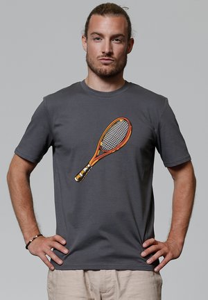 Mann mit zurückgebundenem Haar, der ein graues T-Shirt mit einem großen orangen Tennisschläger-Print trägt und mit den Händen in die Hüften gestützt steht.