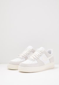 Vita och ljusgrå sneakers med ovandel i mocka och läder, perforerad tå, platta snörningar och texturerad gummisula. Har en Nike-logotyp.