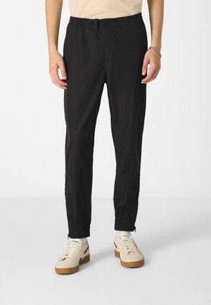 Mand iført sorte tapered bukser med elastiske manchetter og hvide Reebok sneakers, stående mod en ensfarvet hvid baggrund.