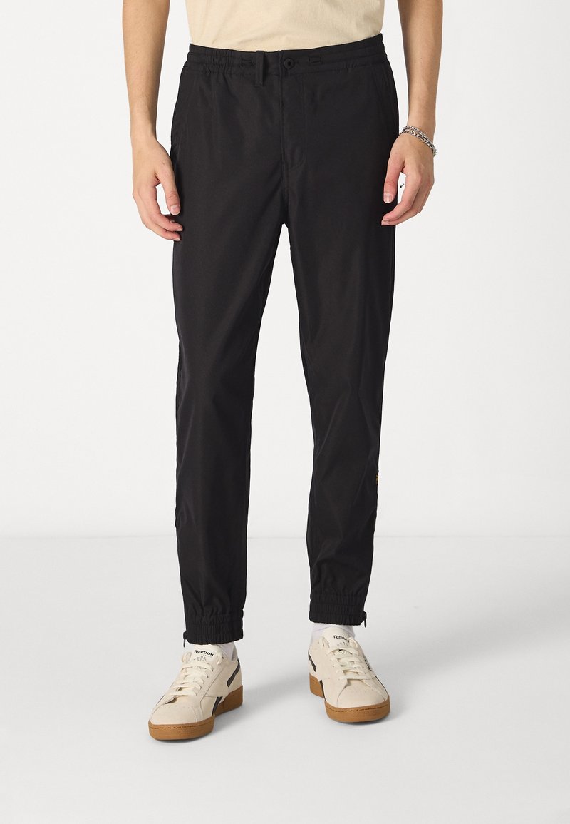 Homme portant un pantalon noir fuselé avec des poignets élastiques et des baskets blanches Reebok, debout devant un fond blanc uni.