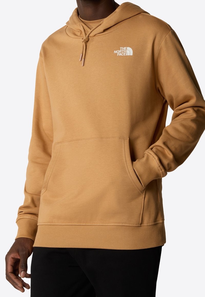 Homme portant un sweat à capuche The North Face couleur camel avec poche avant et capuche à cordon, associé à un pantalon noir, debout sur fond blanc.