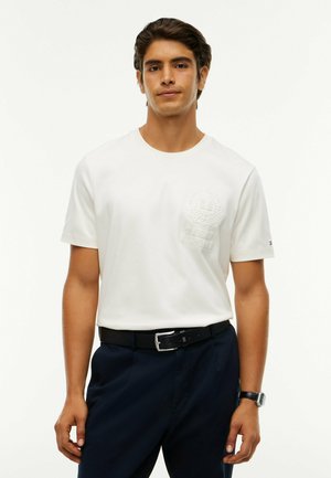 Mand stående iført en hvid kortærmet t-shirt med præglogo, sort bælte, navy bukser og et armbåndsur på venstre håndled.