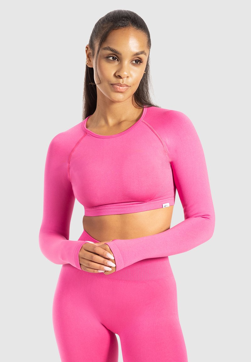 Vrouw met lang haar in een felroze lange mouw crop top en bijpassende high-waisted leggings, staande tegen een effen lichte achtergrond.