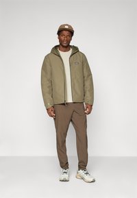 Olijfgroene jas met capuchon, ritssluiting en logo-opdruk. Crème gebreide trui, bruine broek en witte sneakers met grijze accenten.