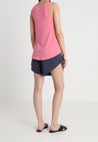 Różowy bawełniany top na ramiączkach bez rękawów, połączony z granatowymi szortami o zaokrąglonym dole. Model nosi czarne klapki.