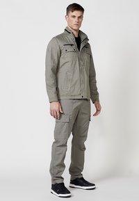 Khaki cargo bunda s viacerými prednými vreckami, vysokým golierom a hladkou textúrou. Kombinovaná s ná matching cargo nohavicami a čiernymi teniskami.