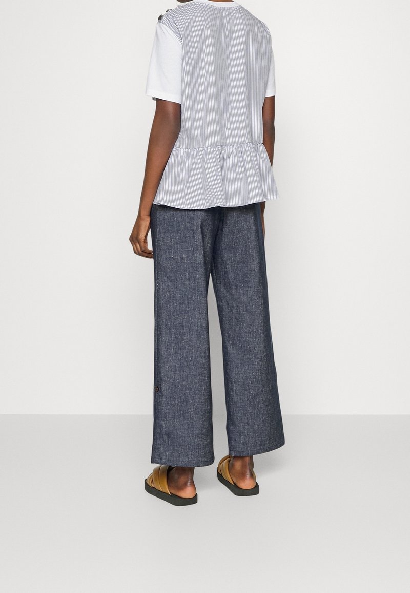 Top senza maniche a righe con design peplum, abbinato a pantaloni in denim scuro larghi e sandali marroni. La vista posteriore mostra il dettaglio dei bottoni sui pantaloni.