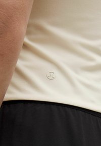 Svijetlosmeđi sportski top s glatkom teksturom, s malim srebrnim logotipom i crnom donjom ivicom. Tkanina je rastezljiva i prilagođena tijelu.