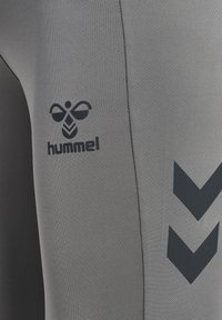 Pantalons de sport gris en tissu texturé avec un logo noir et des motifs en chevrons sur le côté, soulignant un design ajusté.